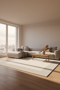 A modern living room featuring a light beige sectional sofa with a matching chaise ottoman, styled with neutral throw pillows and a gray throw blanket. A light wood coffee table sits on a large cream-colored area rug over light hardwood flooring.