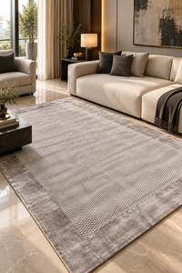 A large rectangular area rug in a warm gray and taupe tone with a textured basketweave pattern and a wide border, shown in a modern living room setting with a beige sofa and marble floor.