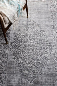 A gray area rug with a distressed medallion pattern, shown in a styled room setting with a wooden chair and a light-colored throw blanket.