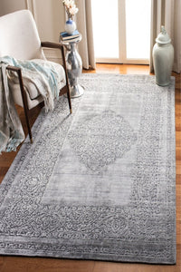 A large rectangular area rug with a faded light gray and white traditional medallion pattern, shown in a styled living room setting with a white armchair, side table, and vase.