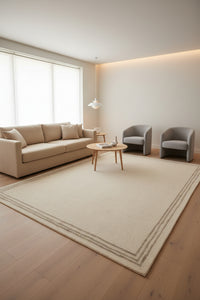 A rectangular area rug in a light beige or cream color, featuring a simple double-line border in a slightly darker beige tone.