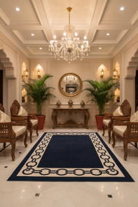 A luxury interior entryway featuring a large navy blue area rug with a cream chain-link border, styled with wooden armchairs, potted palm trees, and a crystal chandelier.