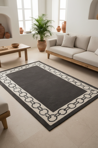 A rectangular area rug with a dark charcoal center and a cream border featuring a repeating interlocking chain pattern, styled in a bright, modern living room next to a light beige sofa.