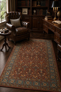 A traditional oriental-style area rug with a red-brown field, blue border, and beige floral motifs, displayed in a classic, dark wood study room next to a leather armchair and desk.
