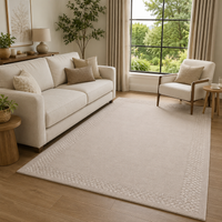 A large rectangular area rug in a warm cream or off-white tone with a subtle textured border, shown in a styled living room setting with a sofa, armchair, wood floor, and large window.